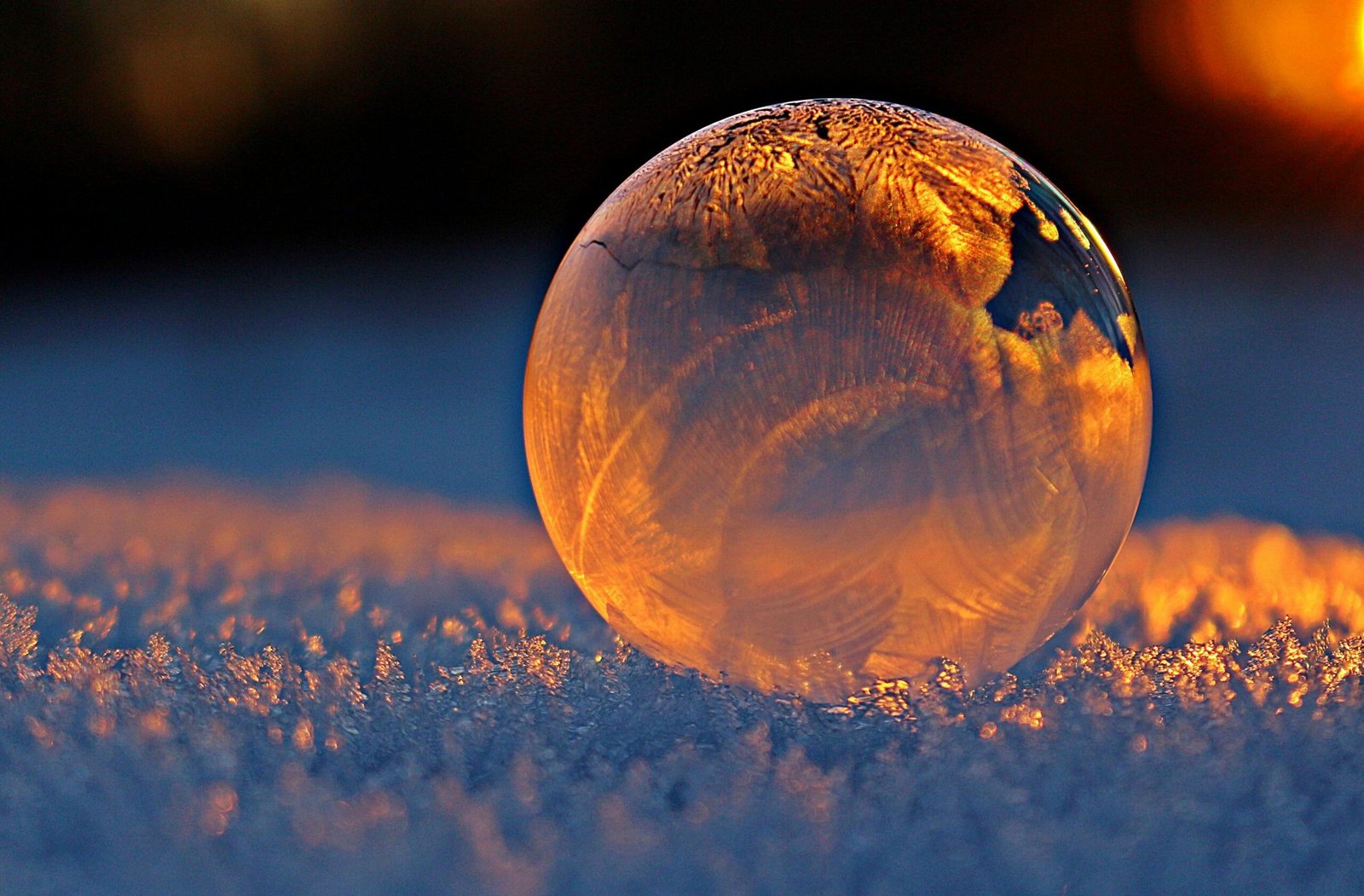 Close-up shot of a frozen bubble with warm reflections resting on a snowy surface at twilight.